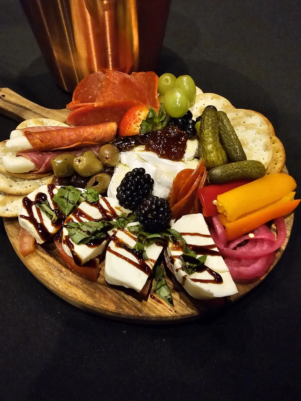 An evening board on a dark table: fresh mozzarella with basil and balsamic, brie, prosciutto, olives, cornichons and peppers.