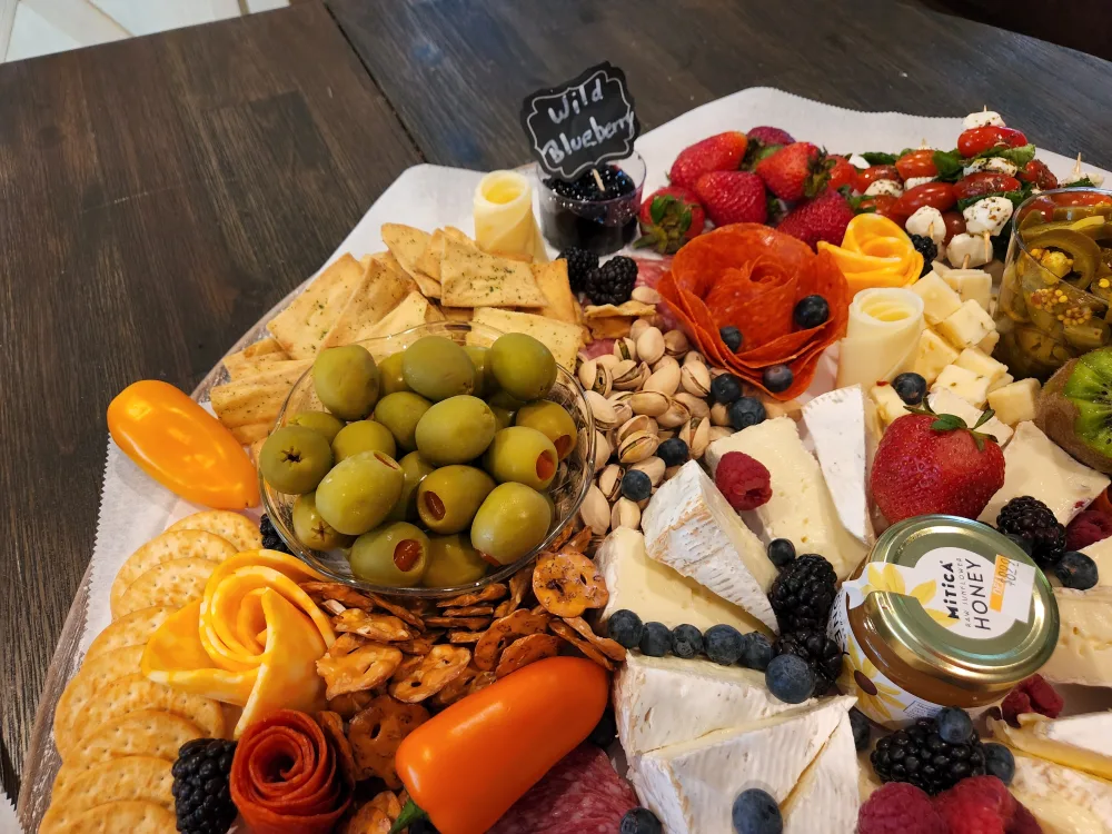 A grazing board seen up close: a bowl of green olives, wedges of brie, pistachios, crackers, cheese roses and fresh berries.
