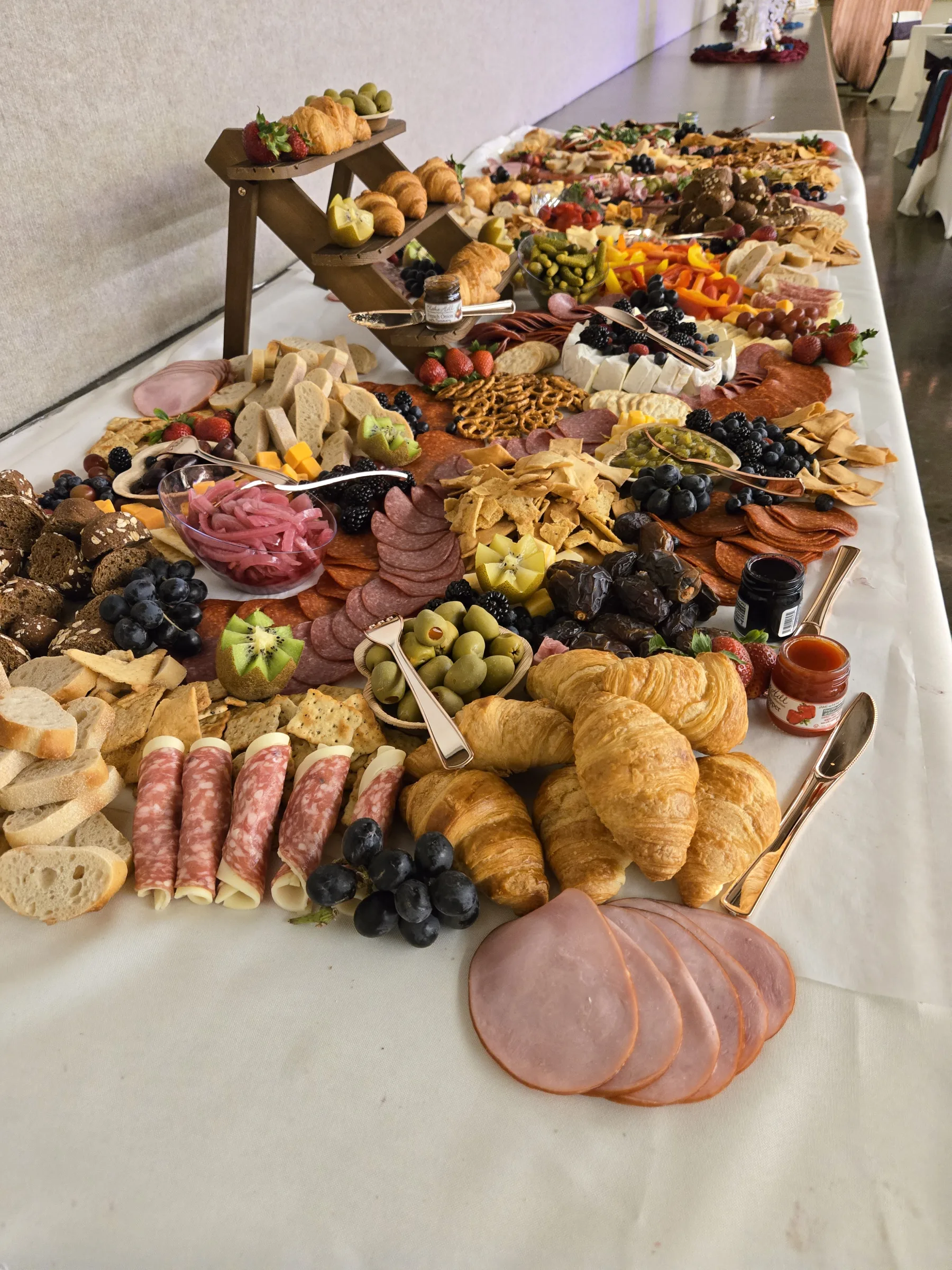 A catered grazing table running the length of a banquet table: cured meats, croissants, cheeses, breads, olives, fruit and preserves.