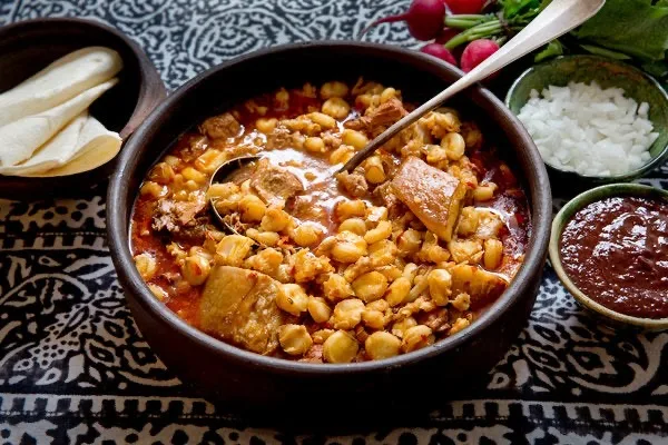 A bowl of posole with hominy and pork, set beside dishes of radish, onion, cheese and red chile for building your own.