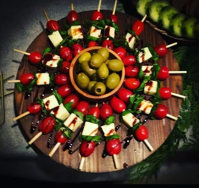 Caprese skewers arranged in a wheel on a round wood board: cherry tomatoes, mozzarella and basil finished with balsamic, set around a bowl of green olives.