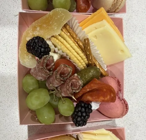An individual graze box: sliced cheeses, salami roses, pepperoni, green grapes, blackberries, crackers, a cornichon and macarons.