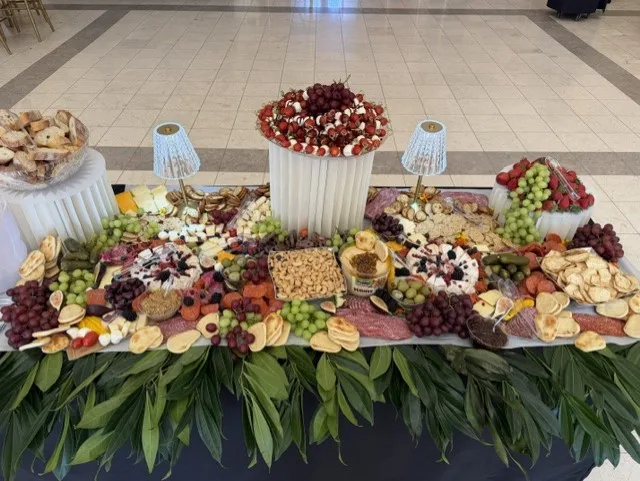 A grazing table set for an event: cheeses, cured meats, crackers, grapes and berries laid the length of the table, a tower of caprese skewers at the center, and greenery running along the front edge.