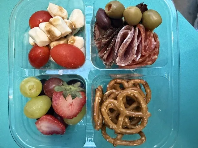 A four-compartment lunch box: mozzarella and cherry tomatoes with balsamic, sliced salami with olives, grapes and strawberries, and pretzels.